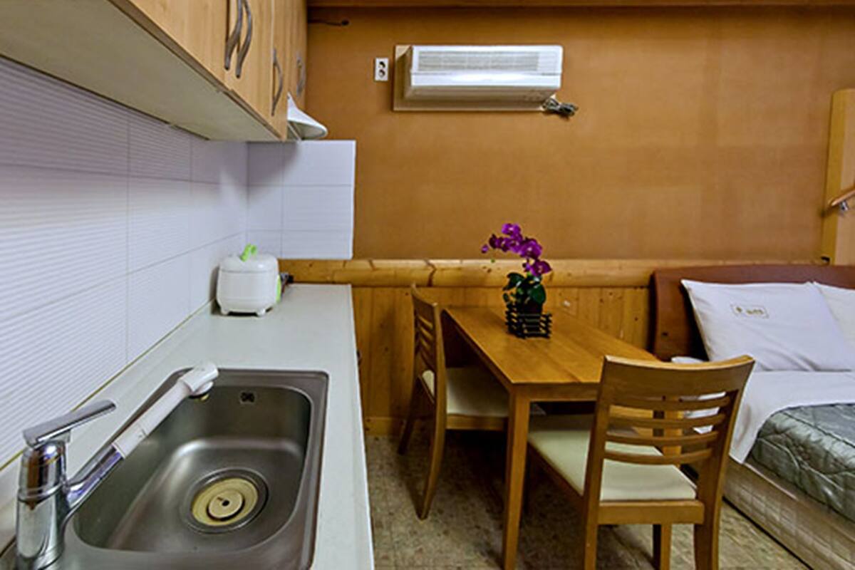 Cocina compacta con pileta, tostadora y una pequeña mesa con sillas. Se observa una ventana con un equipo de aire acondicionado encima de la mesa.