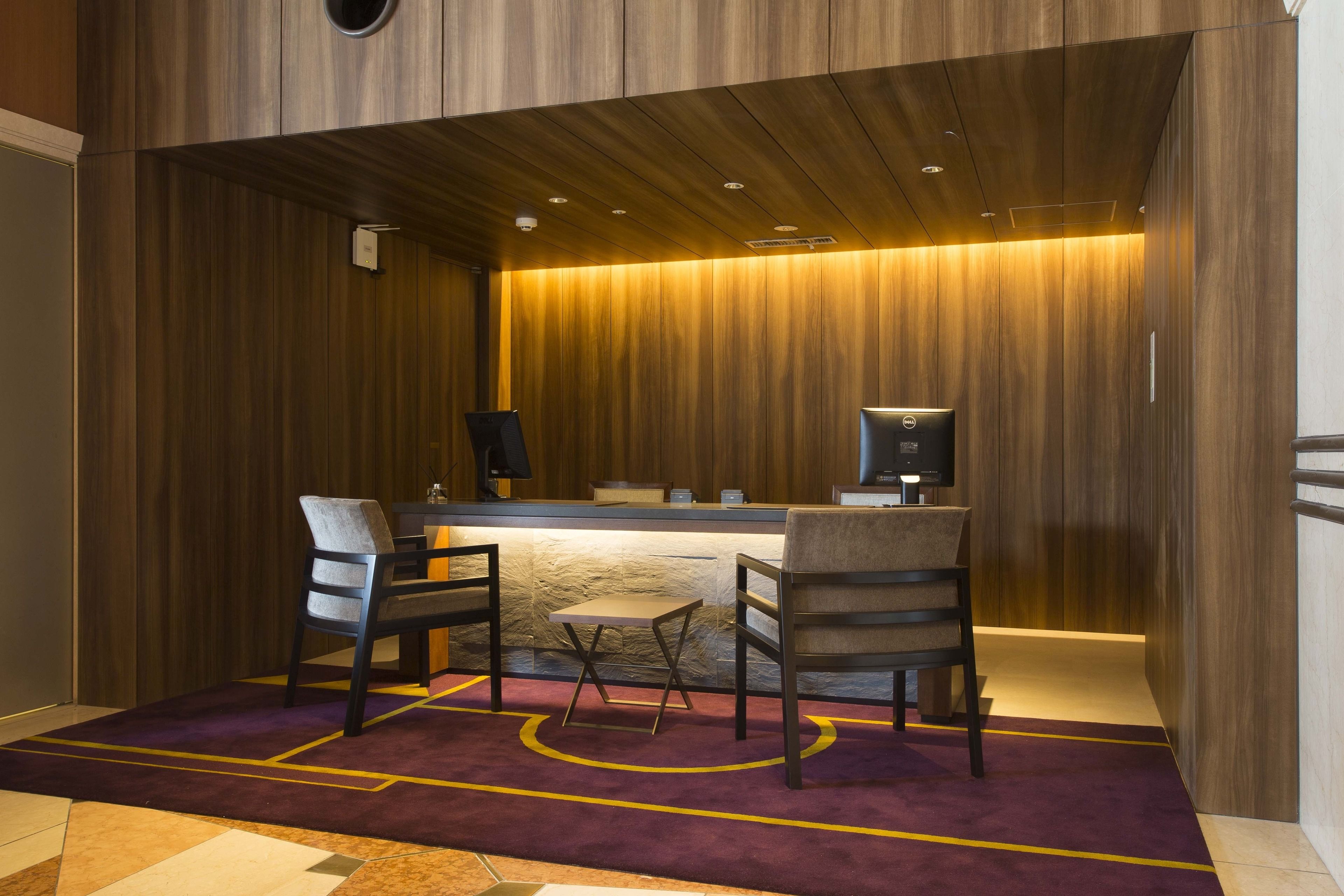 Un hall de hotel con mostrador de recepción, dos sillas, una mesa pequeña y una alfombra morada.