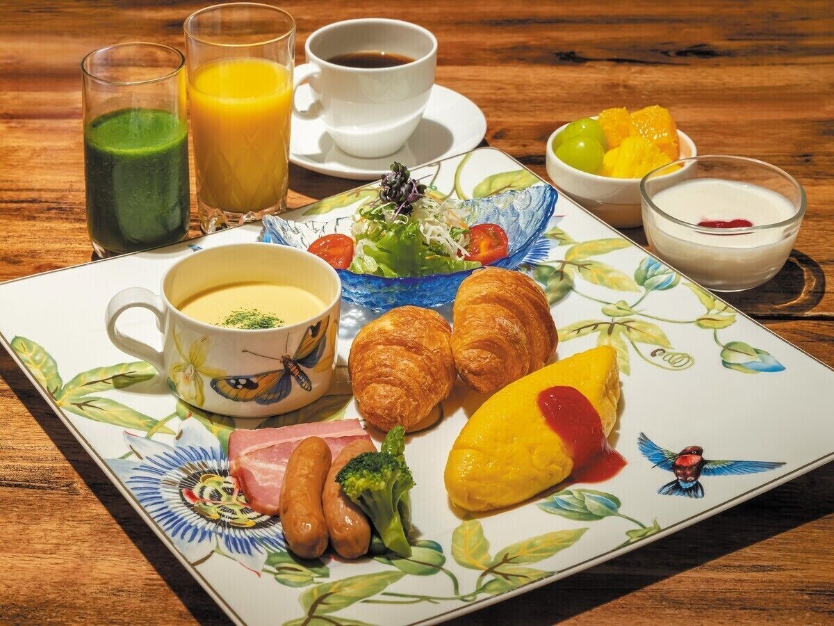 Un plato de desayuno con cruasanes, salchichas, una porción de fruta amarilla, un batido verde, una taza de café y un tazón de yogur.