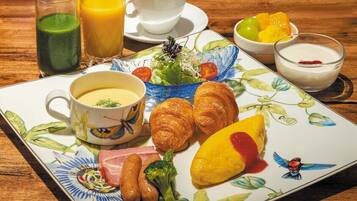 Un plato de desayuno con cruasanes, salchichas, una porción de fruta amarilla, un batido verde, una taza de café y un tazón de yogur.