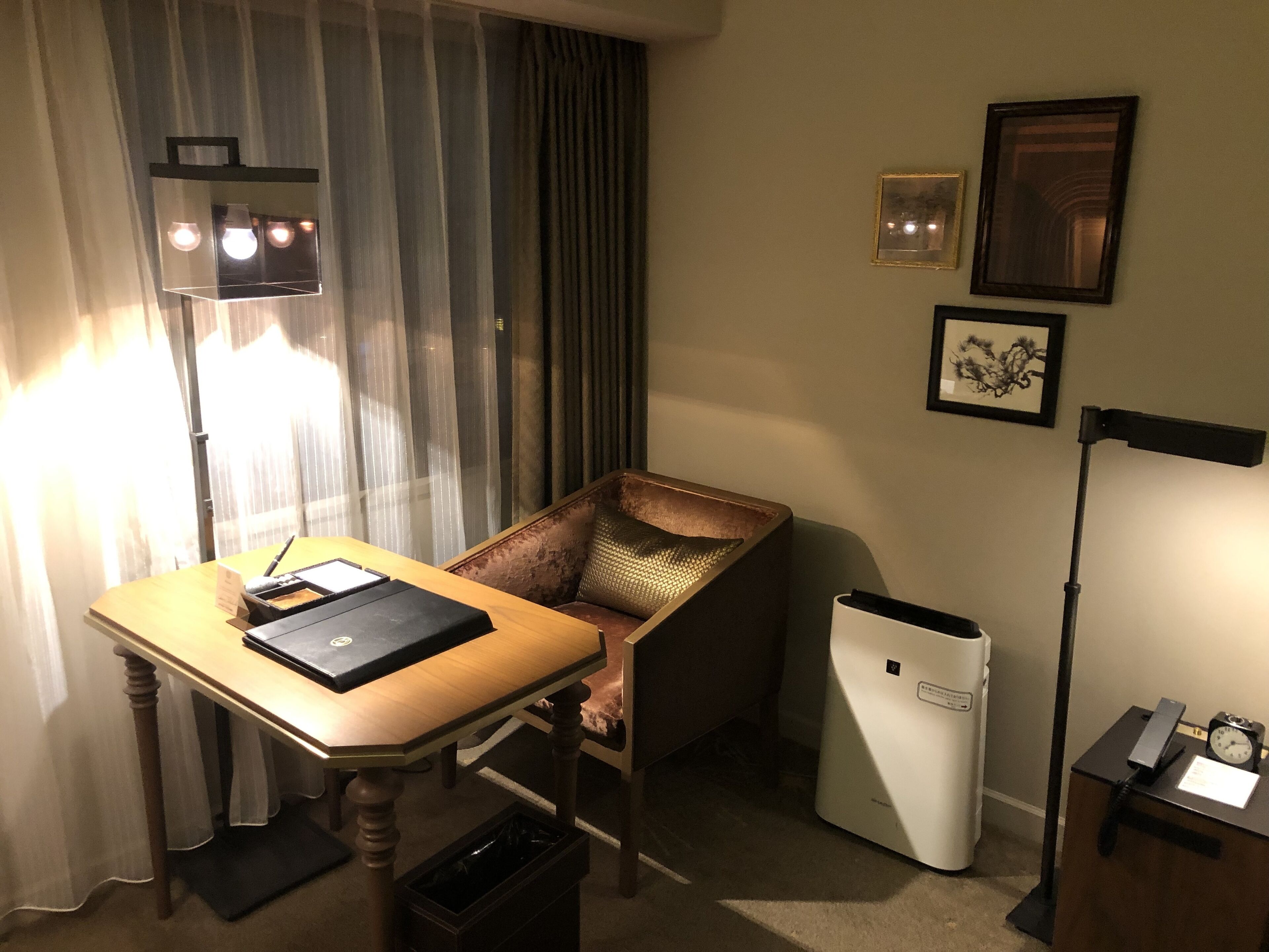 A hotel room with a wooden desk, a chair, a trash bin, a lamp, and a wall-mounted picture.