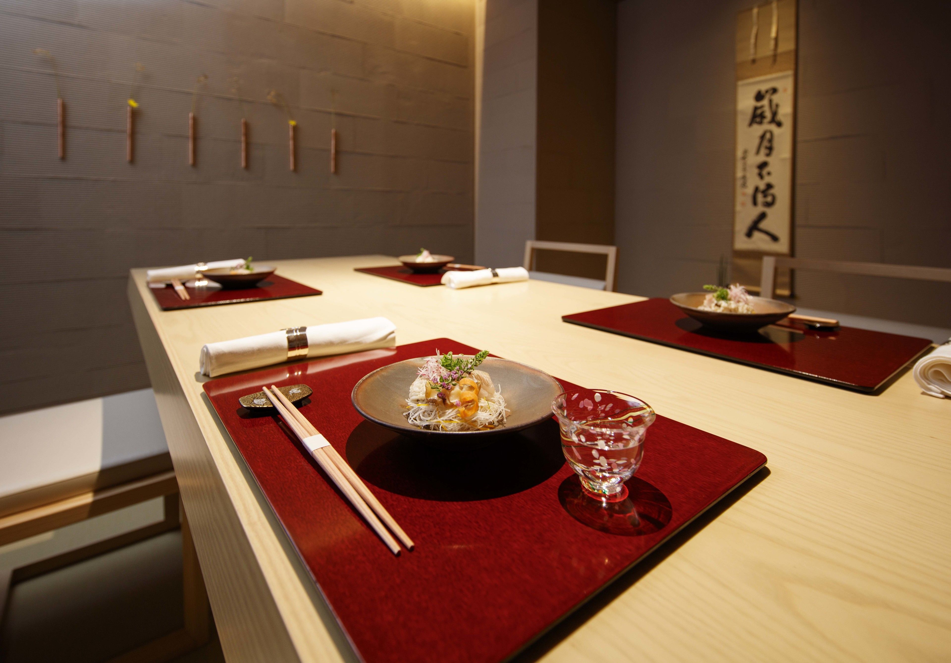 Une table dressée pour deux dans un restaurant, avec un bol de nourriture, des baguettes, un verre d’eau et un set de table rouge.