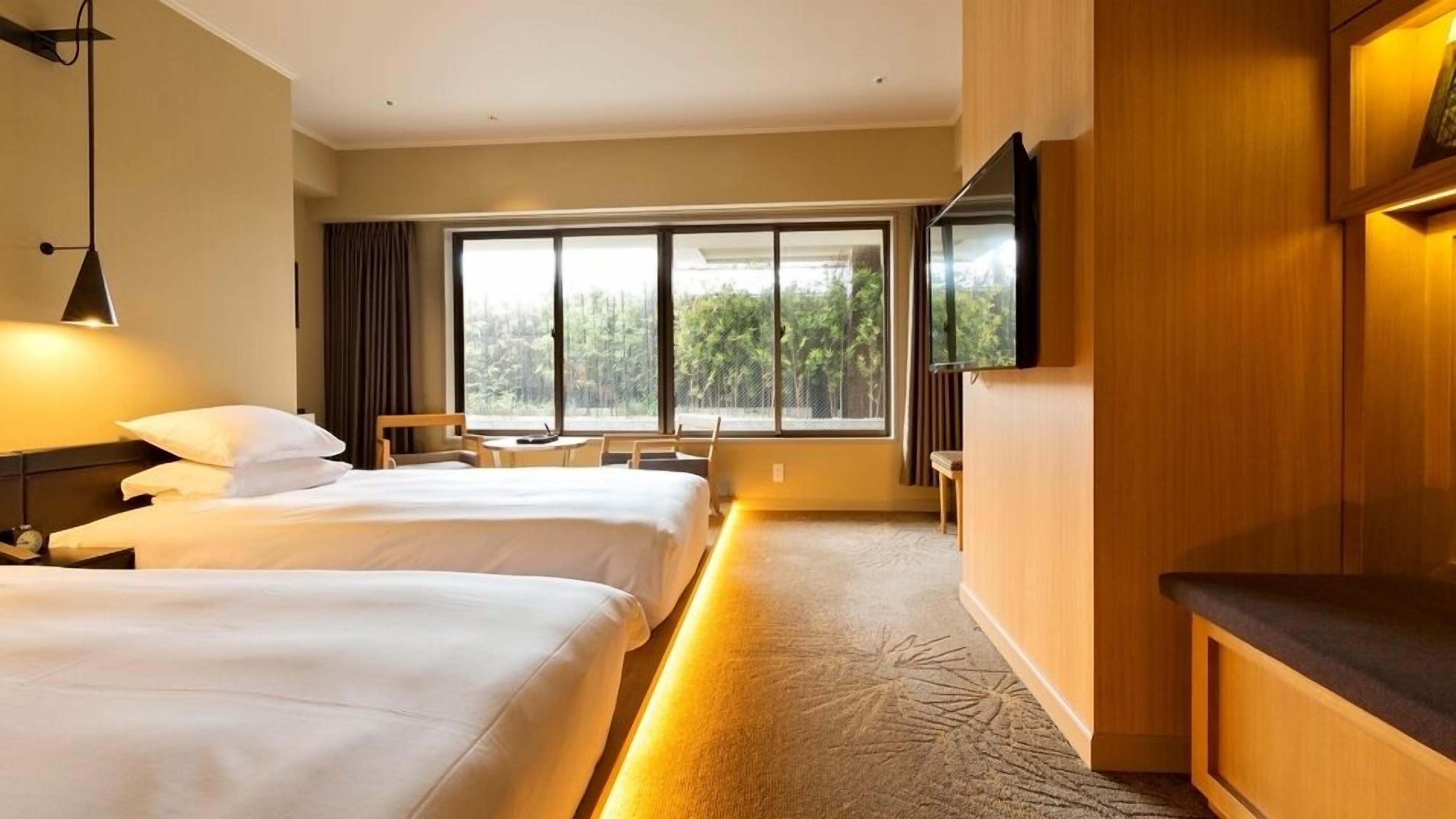 Ein Hotelzimmer mit zwei Betten, einem Fernseher, einem Schreibtisch und einem großen Fenster mit Blick auf Bäume.