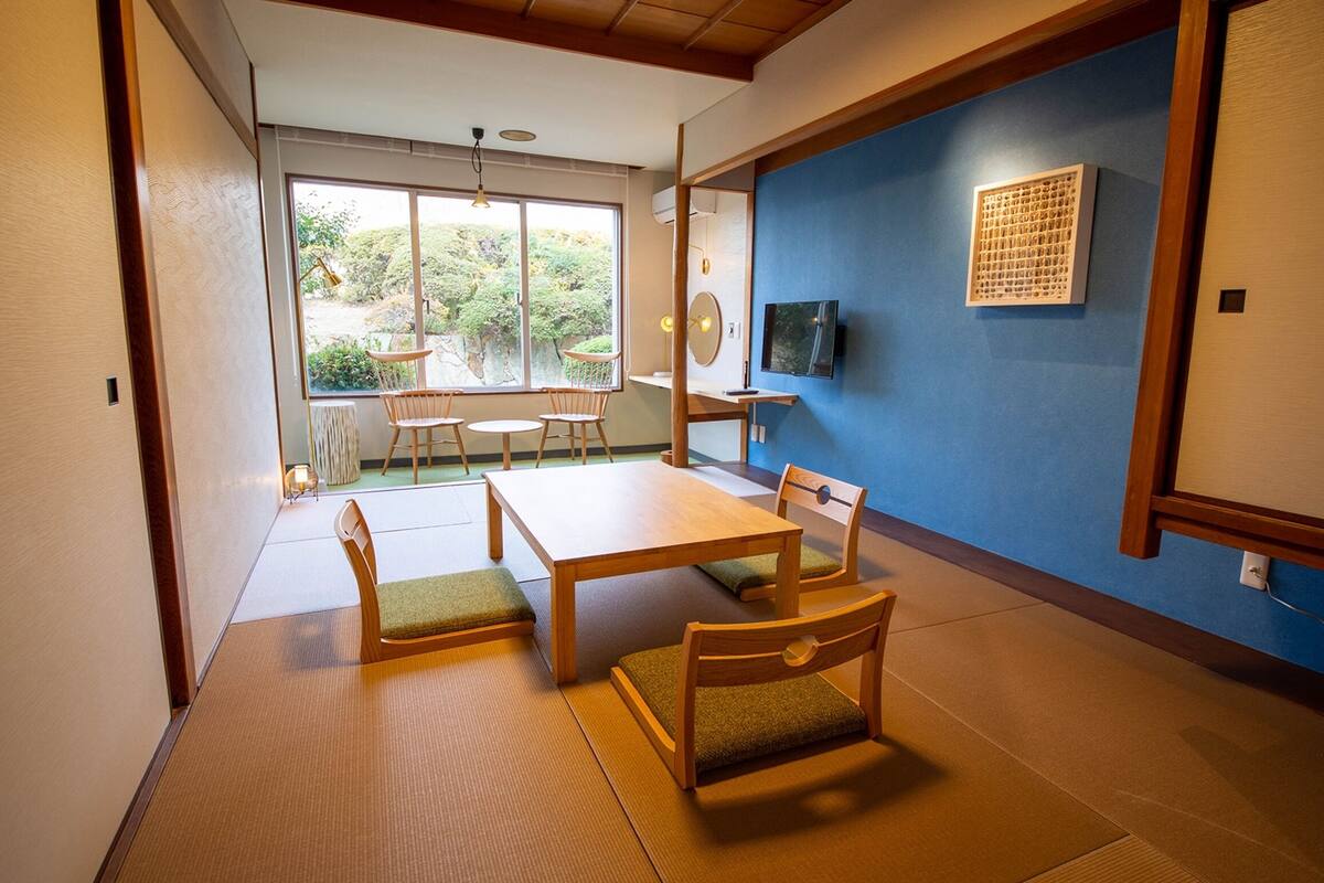 A traditional Japanese room with tatami flooring, a low table with chairs, and a view of greenery outside.