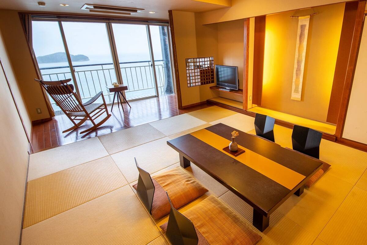 A traditional Japanese-style room with tatami flooring, a low table with cushions, a television, and a sliding door leading to a balcony with a view of the sea.