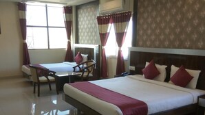 Une chambre d’hôtel avec un grand lit, un lit plus petit, un bureau et une chaise. Il y a des rideaux, un ventilateur de plafond et une fenêtre.