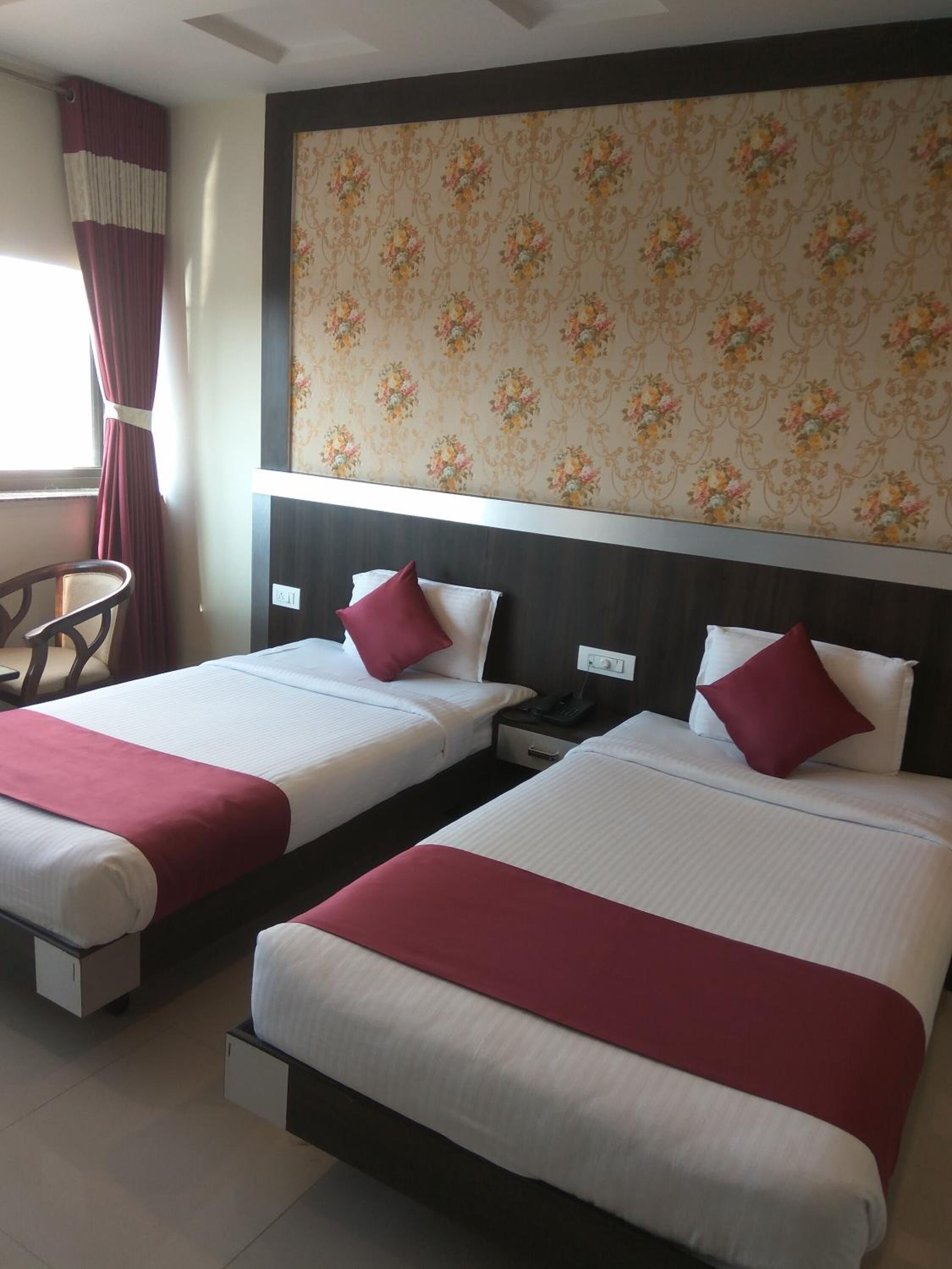 A hotel room with two beds, each with white bedding and red accents, a floral-patterned headboard, and a chair by the window.