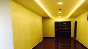 Un couloir aux murs jaunes et au sol brun brillant, qui mène à une porte noire sur laquelle est apposée une enseigne.