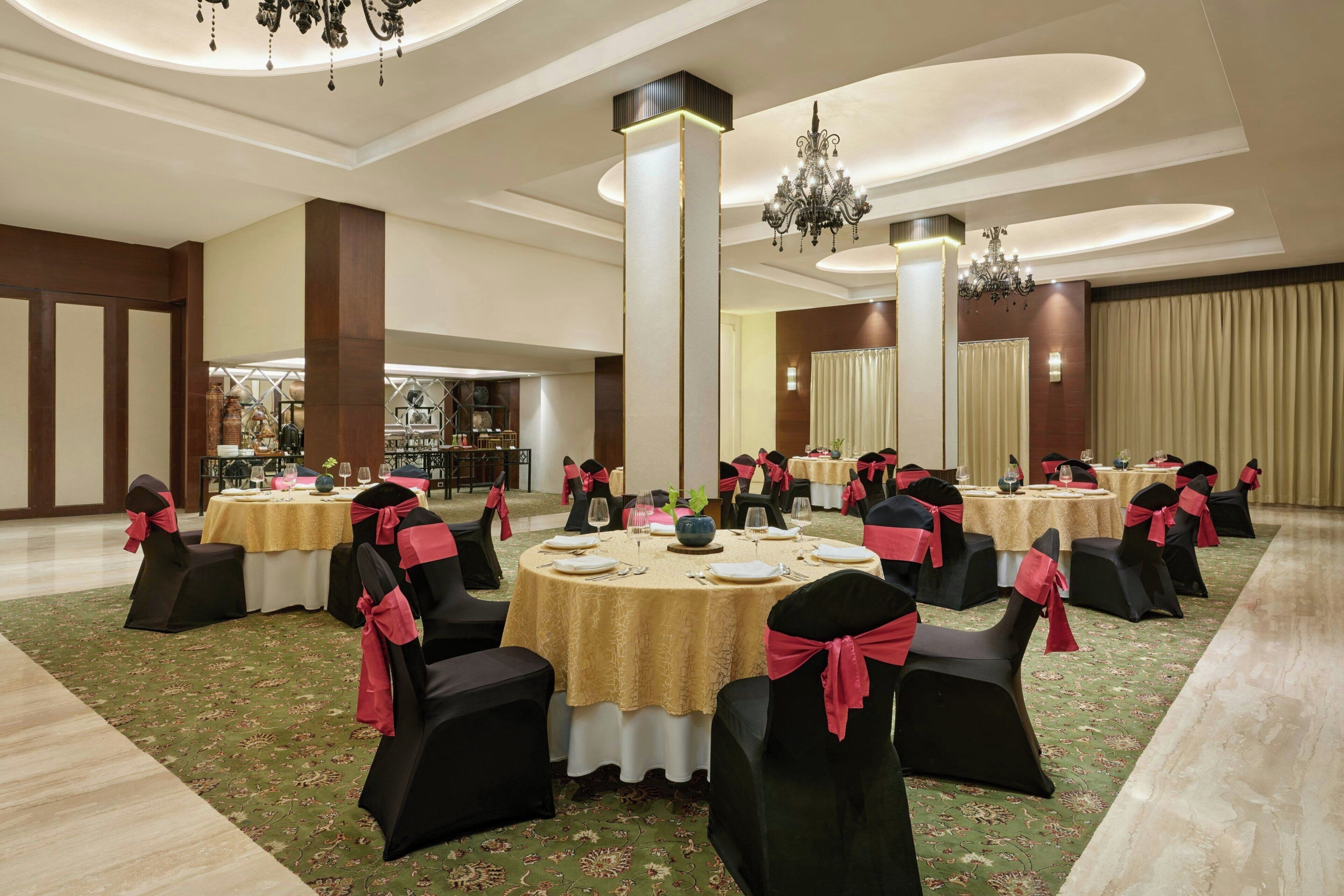 A banquet hall with round tables, black chairs with red sashes, chandeliers, and a high ceiling.