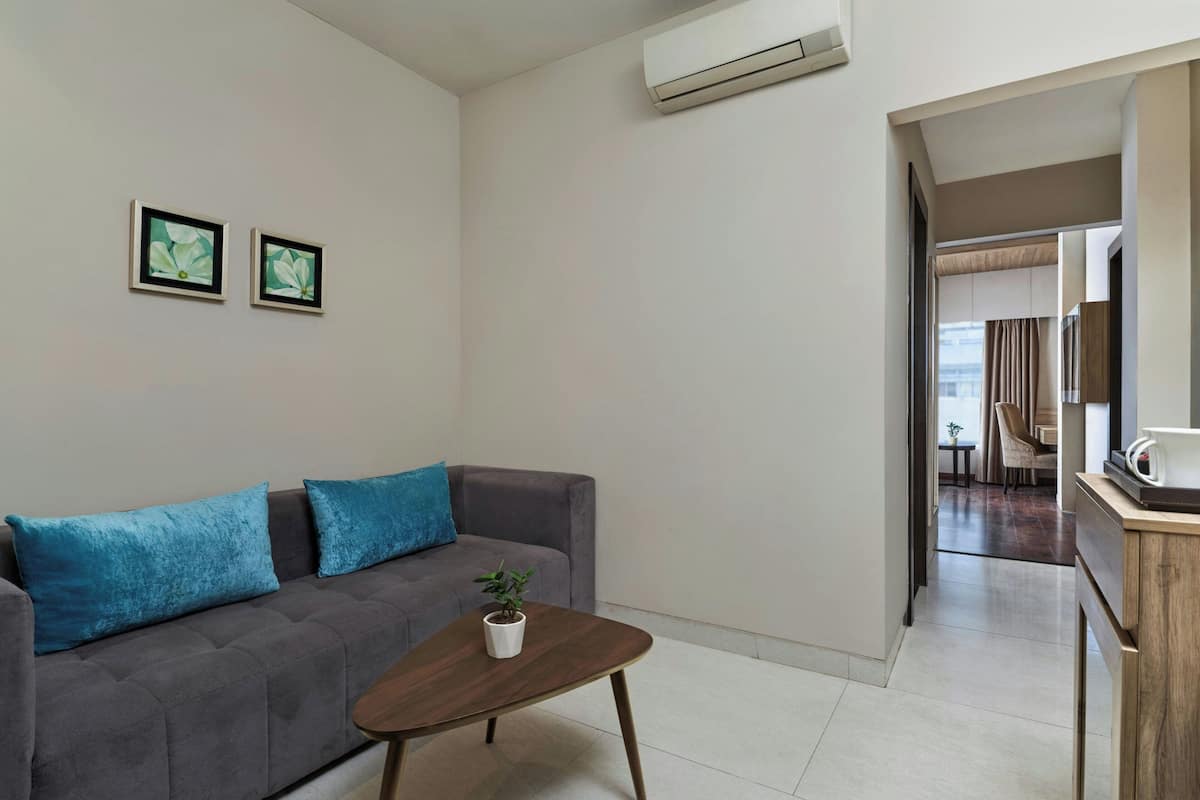 Una sala de estar moderna con un sofá gris, cojines azules, una mesita de centro de madera y una planta en maceta. Hay dos cuadros en la pared y un equipo de aire acondicionado en la parte superior.