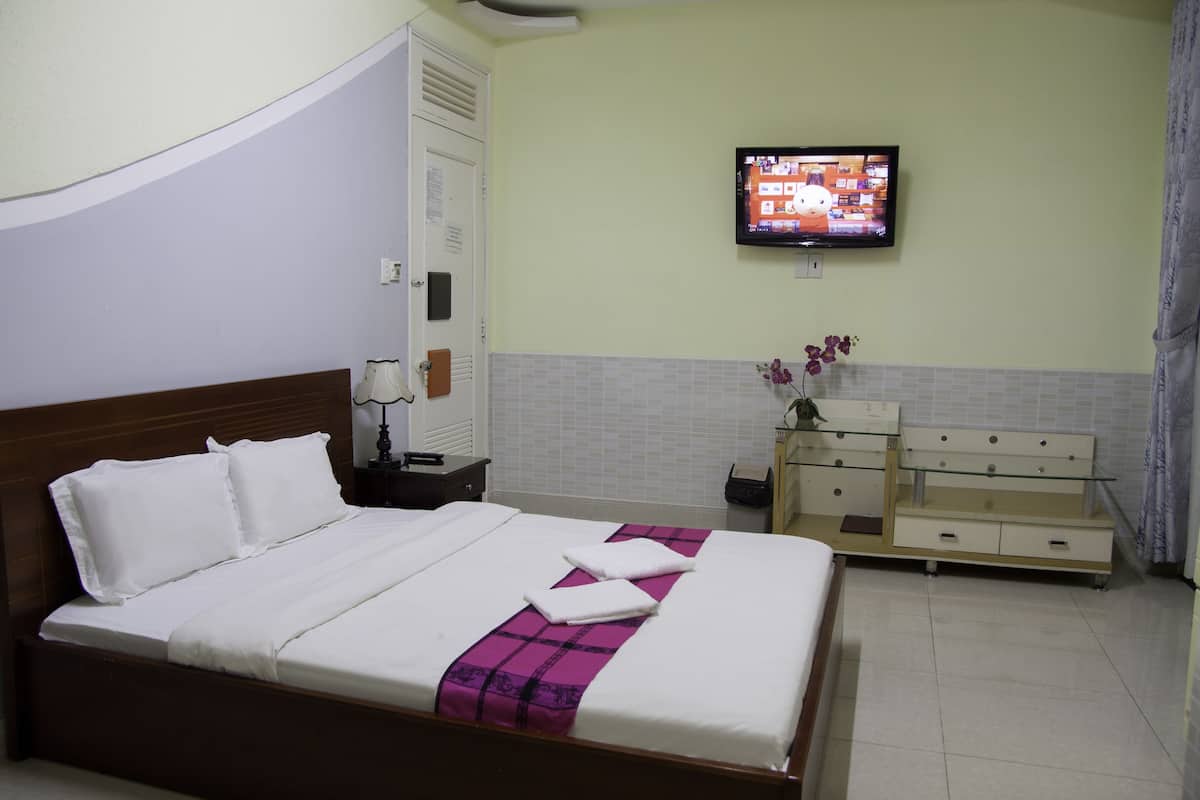 A neatly made bed with white and purple bedding, a television on the wall, and a dresser with drawers.