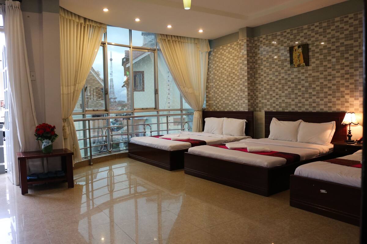 A hotel room with two beds, a balcony with a railing, a small table with a vase of flowers, and a checkered wall pattern.