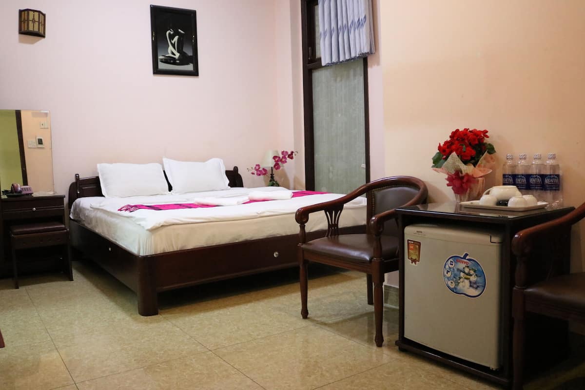 A hotel room with a bed, a desk with a chair, a small refrigerator, and a window with curtains.