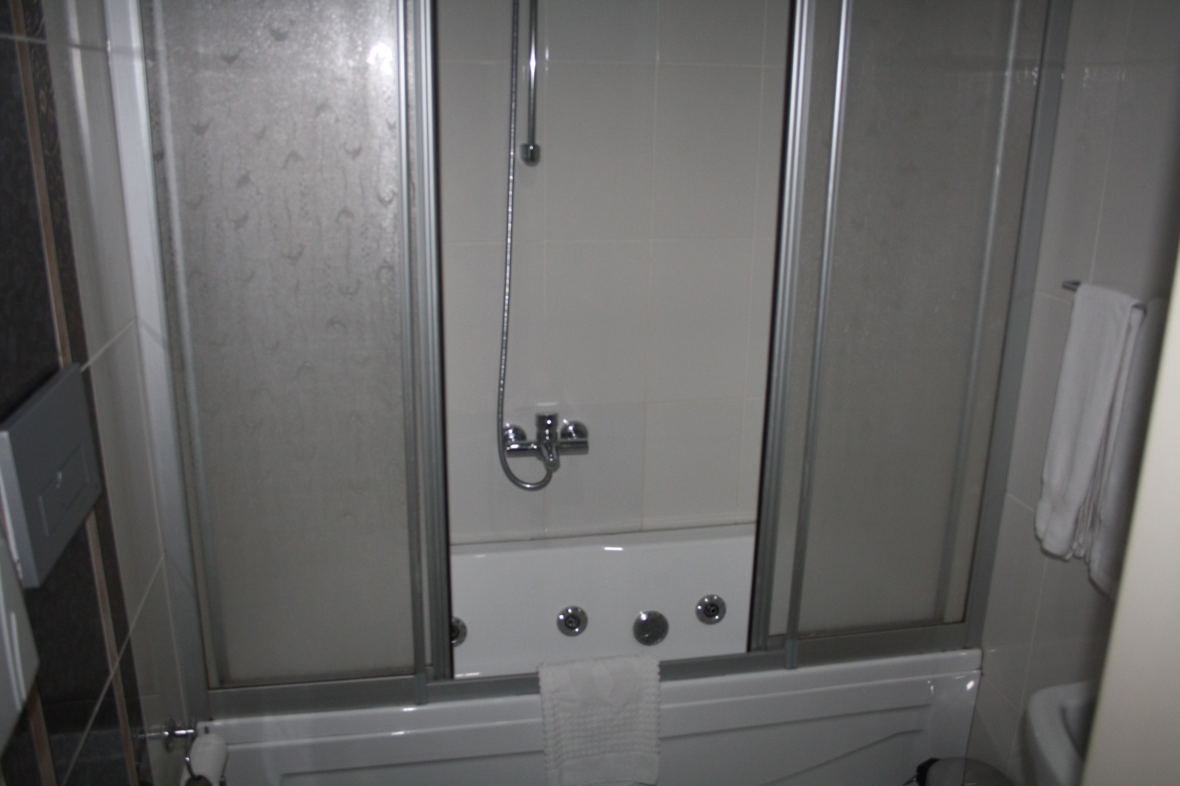 Une cabine de douche fermée par des parois en verre, dotée d’un pommeau de douche à main, d’une serviette accrochée à la porte et d’un porte-rouleau de papier toilette visible.