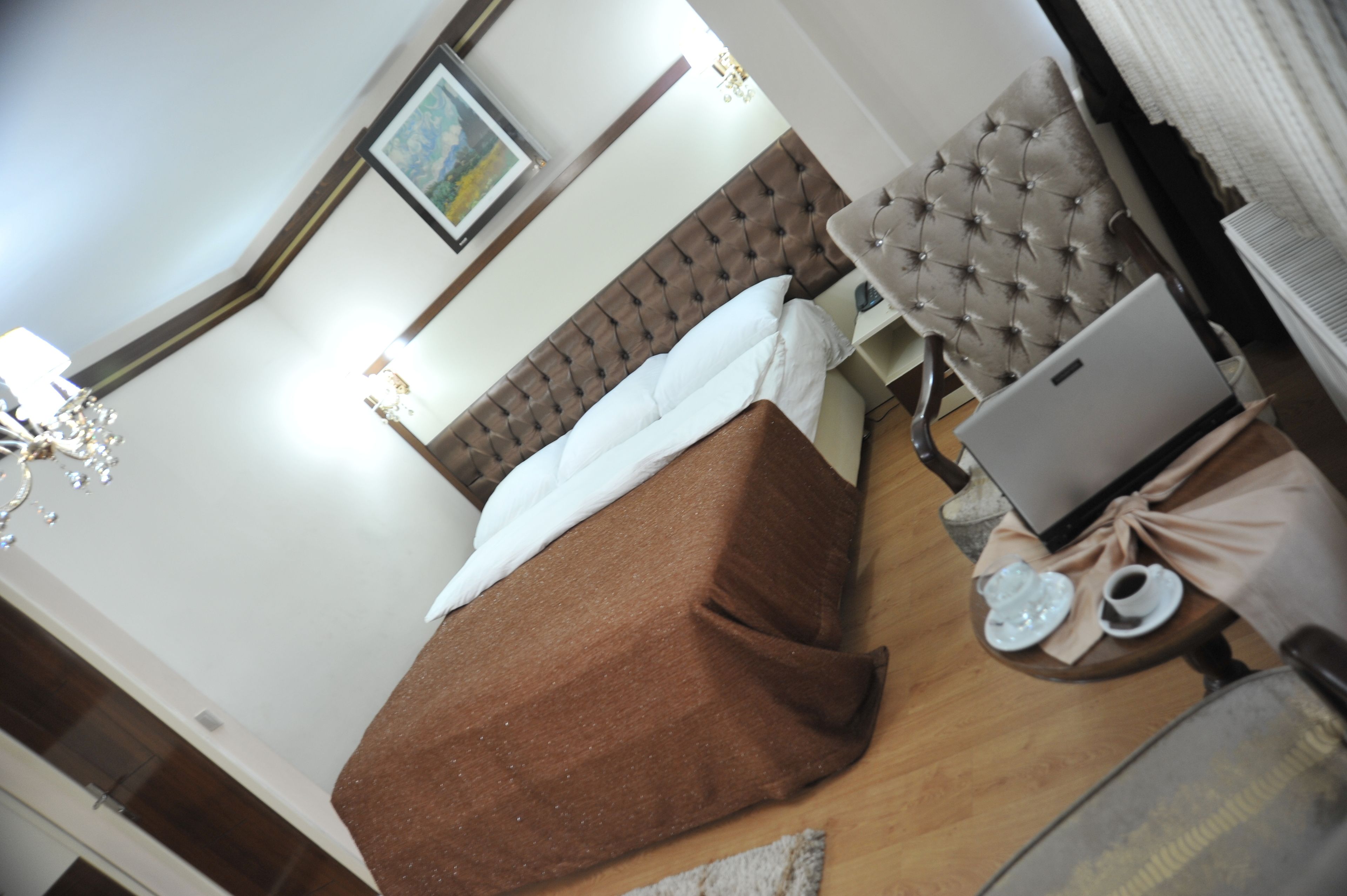 A bedroom with a bed, a laptop, a chair, and a table with a teapot and cup.