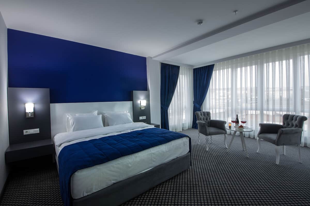 A modern hotel room with a large bed, two bedside tables, a small round table with a chair, and a view of the city through large windows.