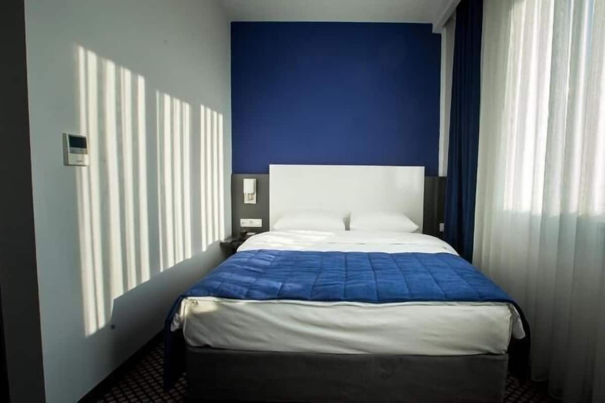 A hotel room with a bed, a nightstand, a wall-mounted phone, and a window with curtains.