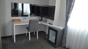 Habitación de hotel con escritorio, dos monitores, una silla, un refrigerador pequeño, cafetera y una botella de agua.