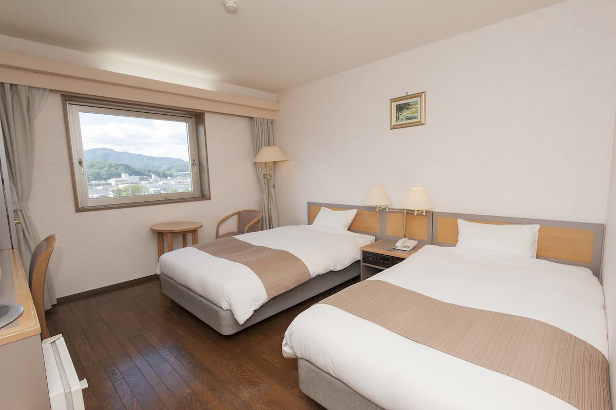 A hotel room with two beds, a desk, a chair, a small table, a lamp, and a window with a view of mountains.