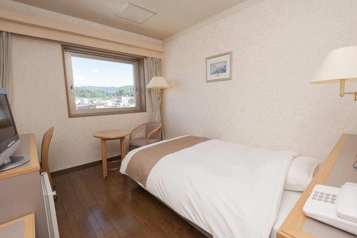 A hotel room with a bed, a desk with a computer, a chair, a window with a view, and a lamp.