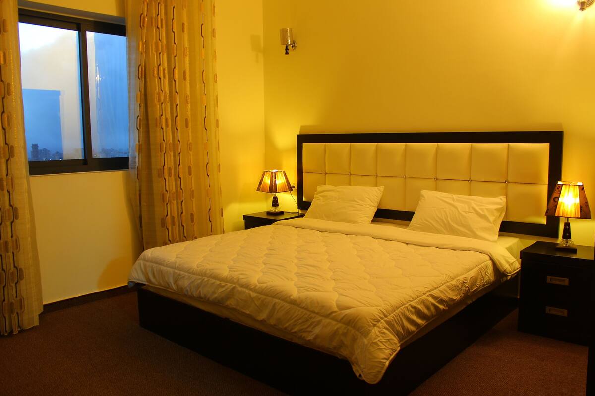 Habitación de hotel con una cama grande, dos mesitas de noche con lámparas, una ventana con cortinas y vistas a la ciudad.