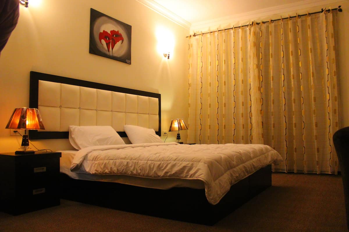 Una habitación de hotel con una cama grande, dos mesitas de noche con lámparas, un cabecero, una obra de arte en la pared y una cortina.