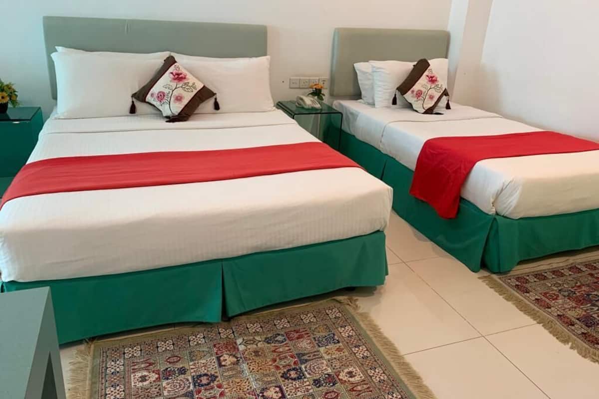 Two beds with green bedspreads, white linens, and red blankets. There are two nightstands with lamps and a small table with a vase of flowers.