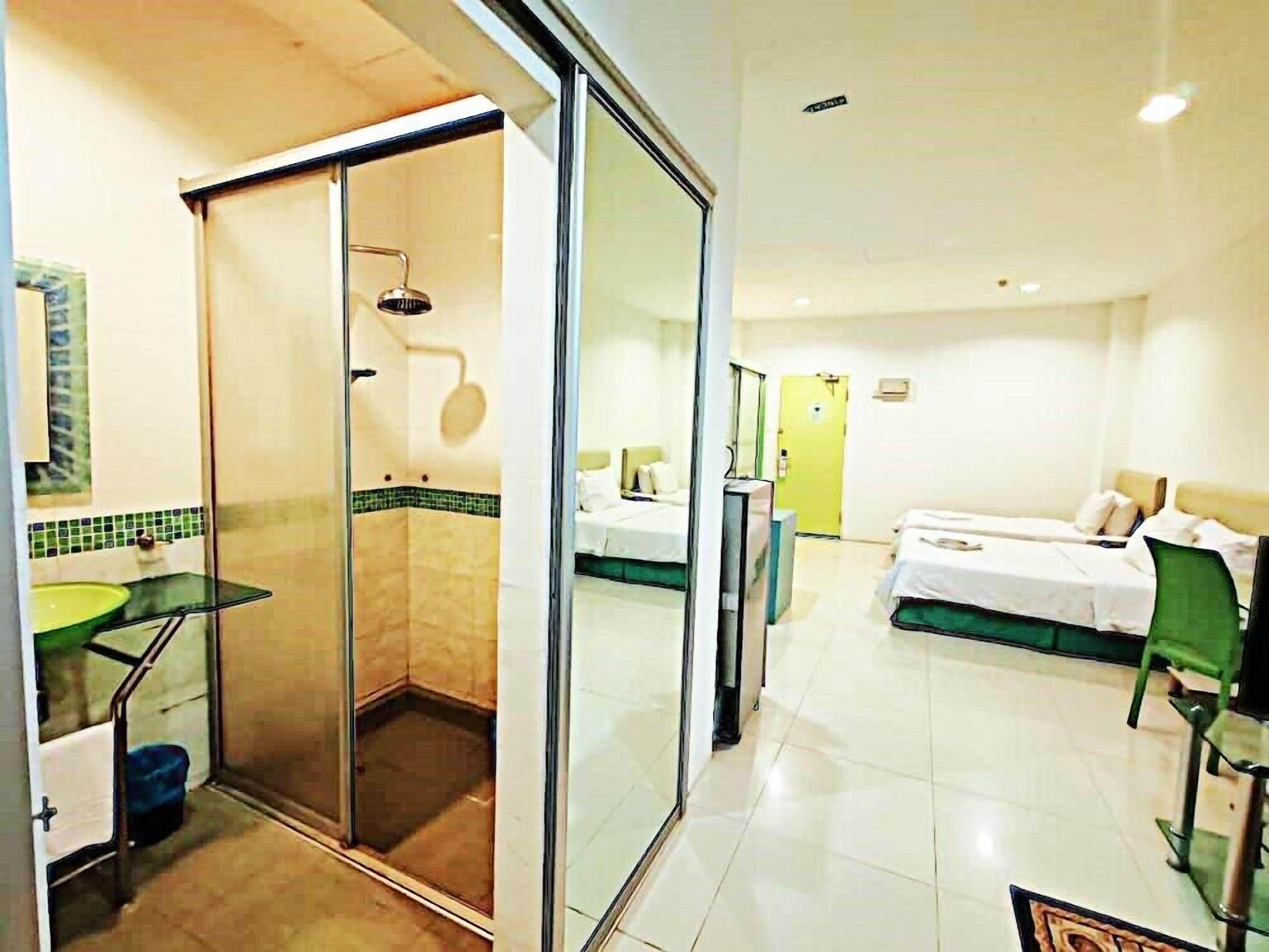 A hotel room with a glass shower enclosure, a bed with green bedding, a bedside table, and a chair.