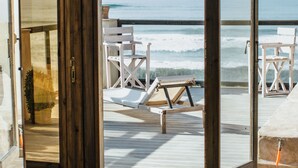 Een kamer met een houten vloer, een bed en een grote schuifdeur naar een balkon met uitzicht op de oceaan en strandstoelen.