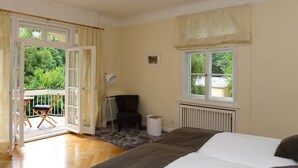 A bedroom with a bed, a chair, a small table, a radiator, and a window with a view of greenery. - Residenz am englischen Garten (Munich)