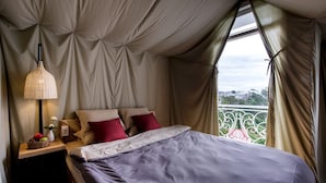 Un dormitorio con cama, almohadas, una mesita de noche con lámpara y vistas a la vegetación por una ventana.