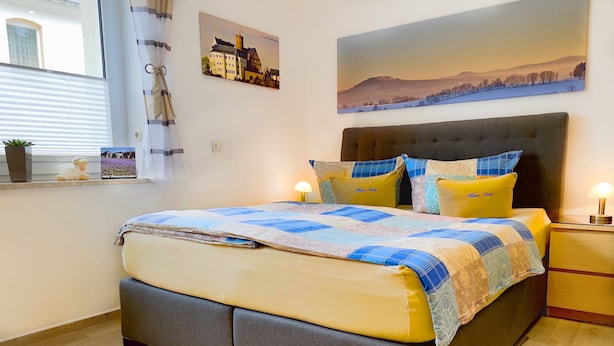 Quarto com cama, mesa de cabeceira, janela com cortinas e quadros nas paredes.