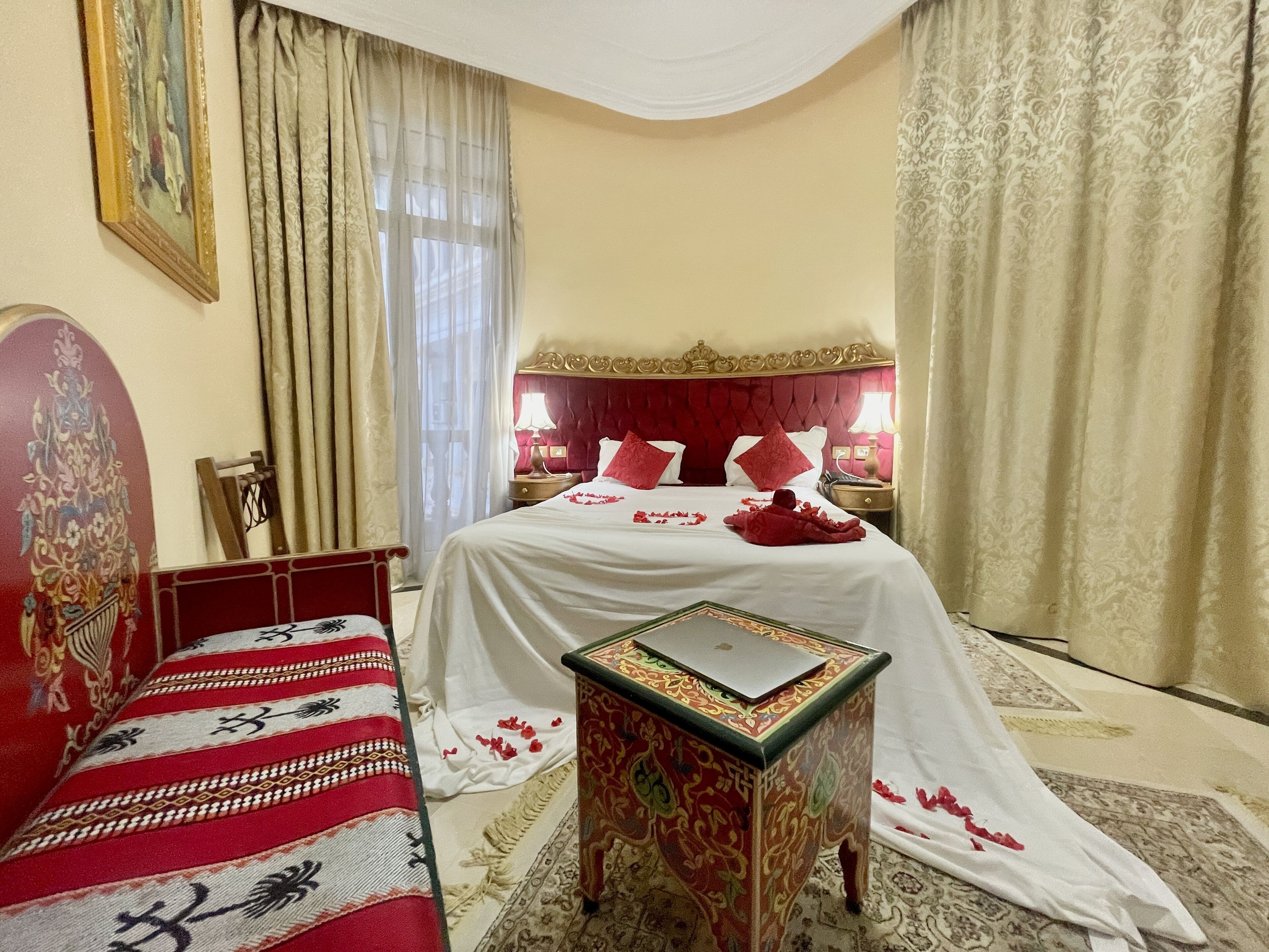 A hotel room with a bed, a small table, a nightstand, a chair, and a painting on the wall.