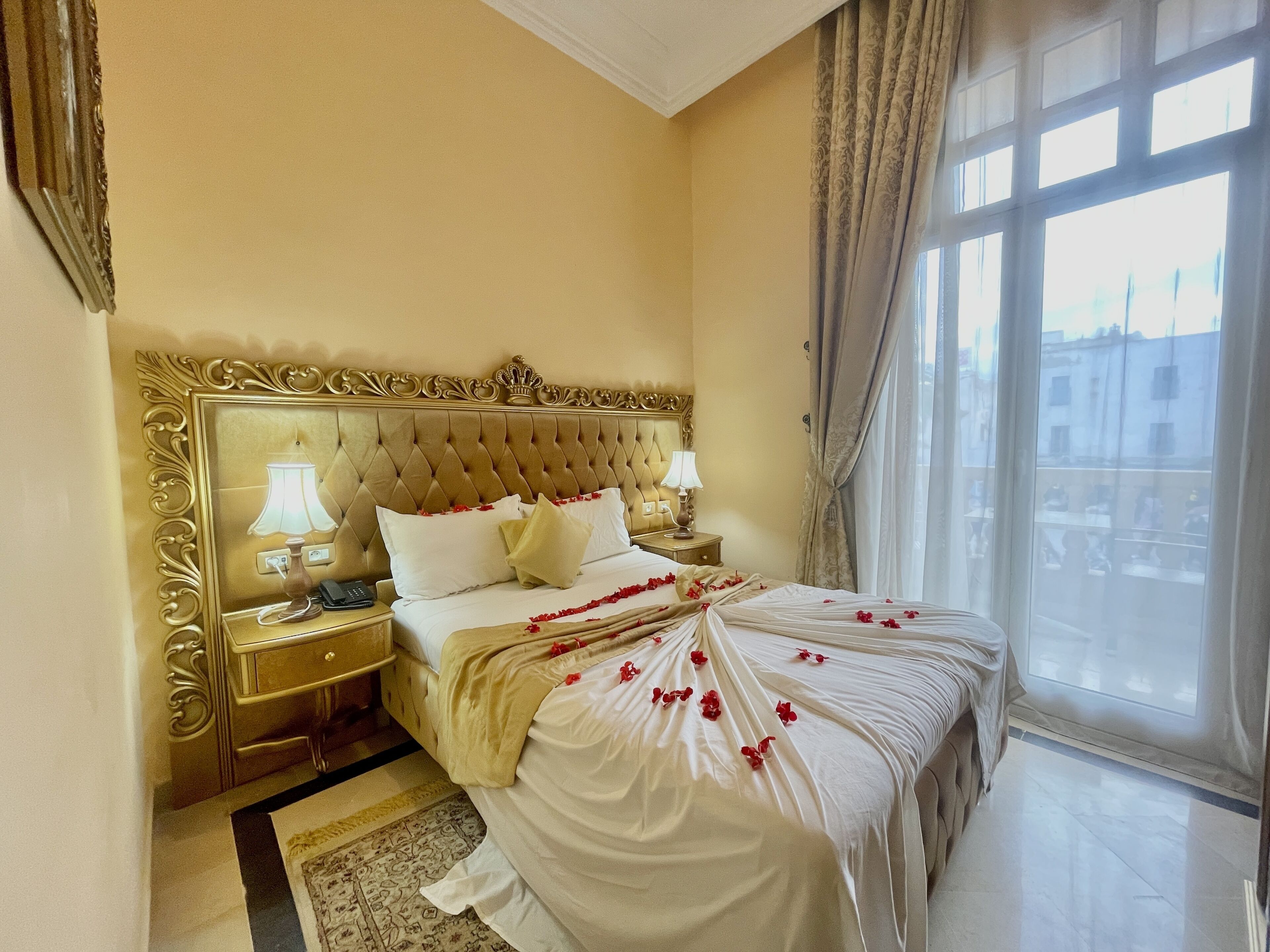 A hotel room with a large bed, ornate headboard, two bedside tables with lamps, and a view of the city through large windows.
