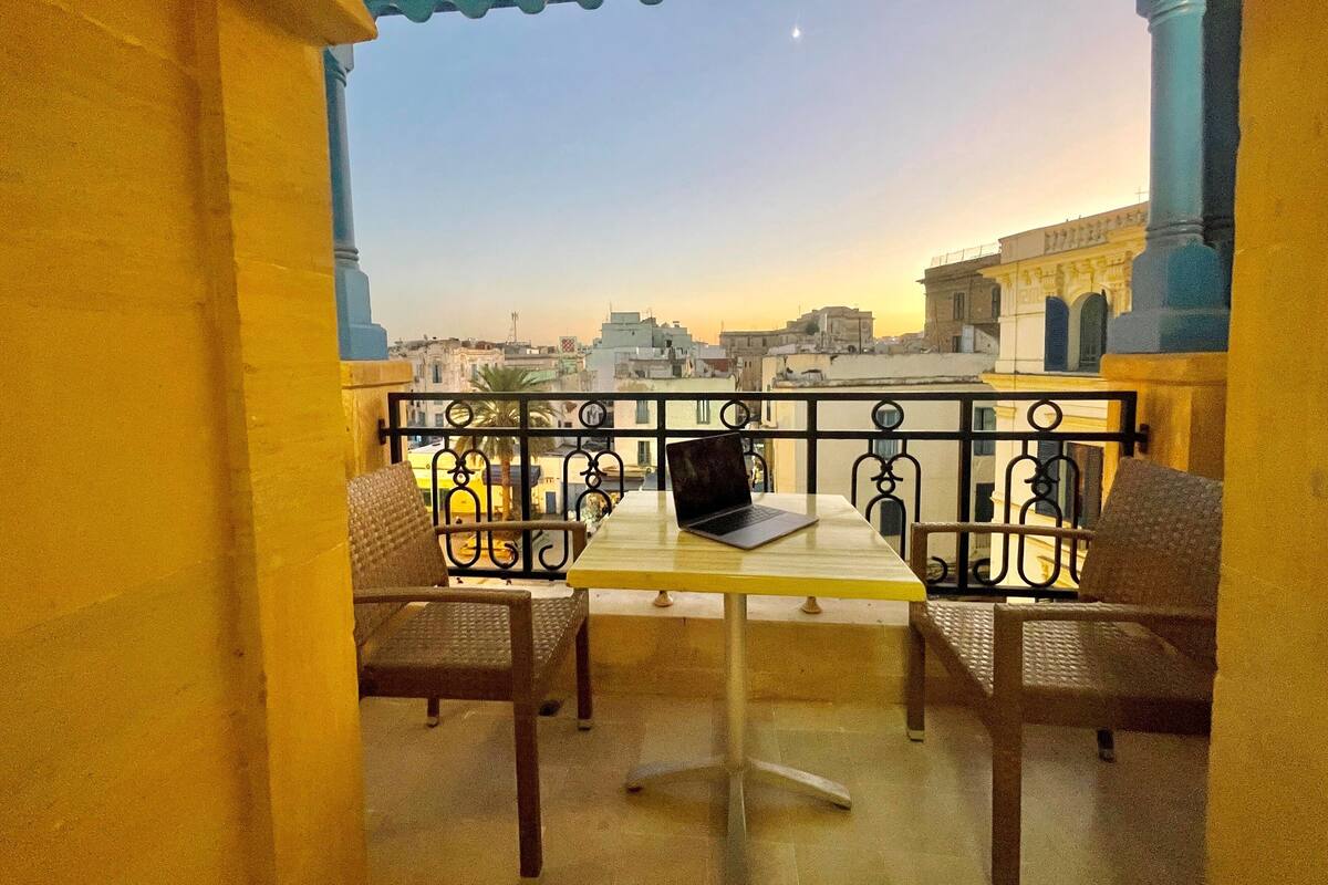 A balcony with a table and two chairs, overlooking a cityscape with buildings and a clear sky.