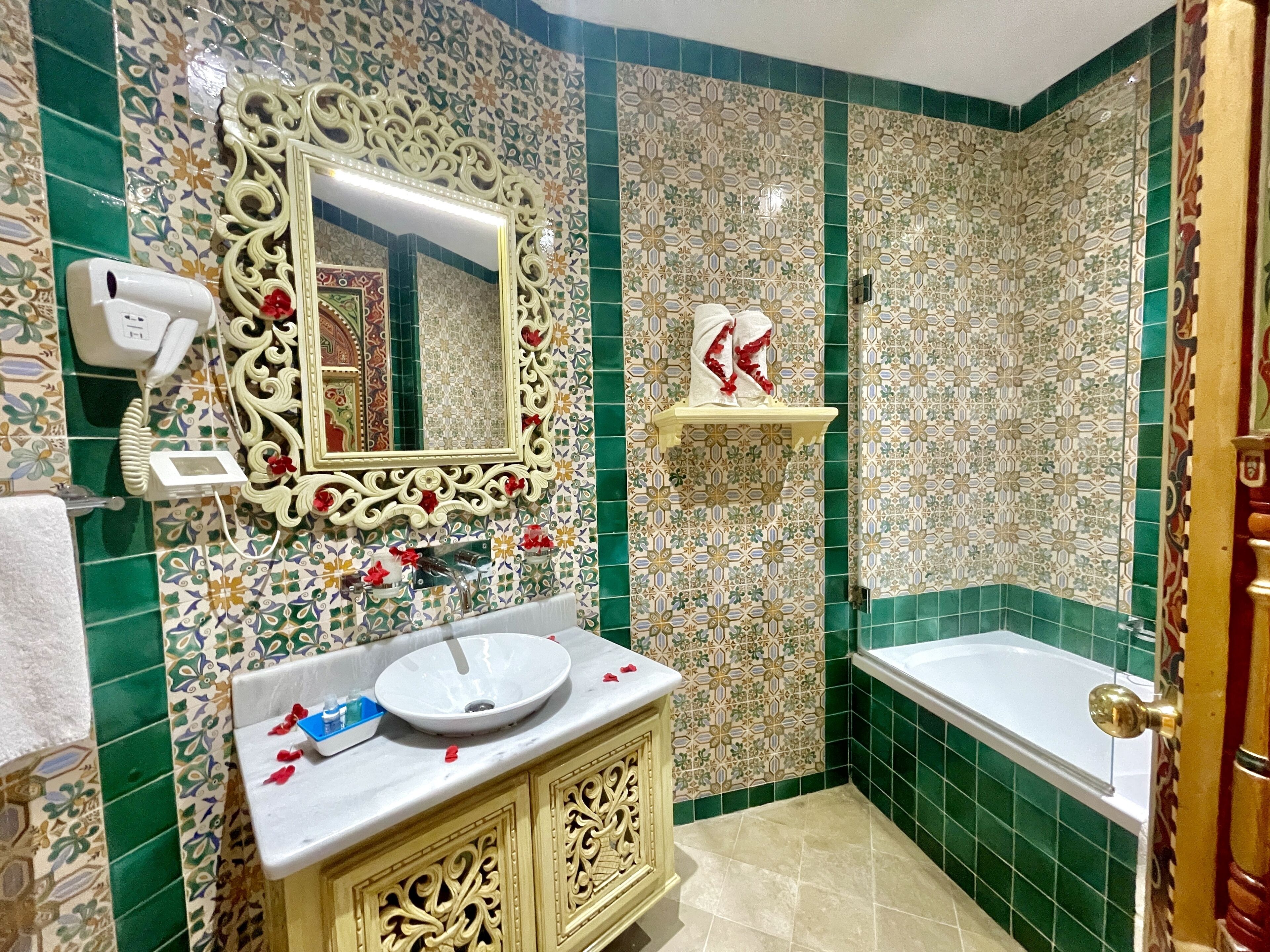 A bathroom with a white sink, ornate gold cabinetry, a large mirror, and a green-tiled shower area.