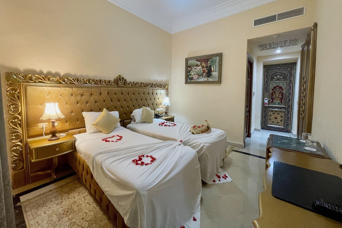 A hotel room with two beds, a nightstand, a lamp, a painting, and a door with intricate designs.