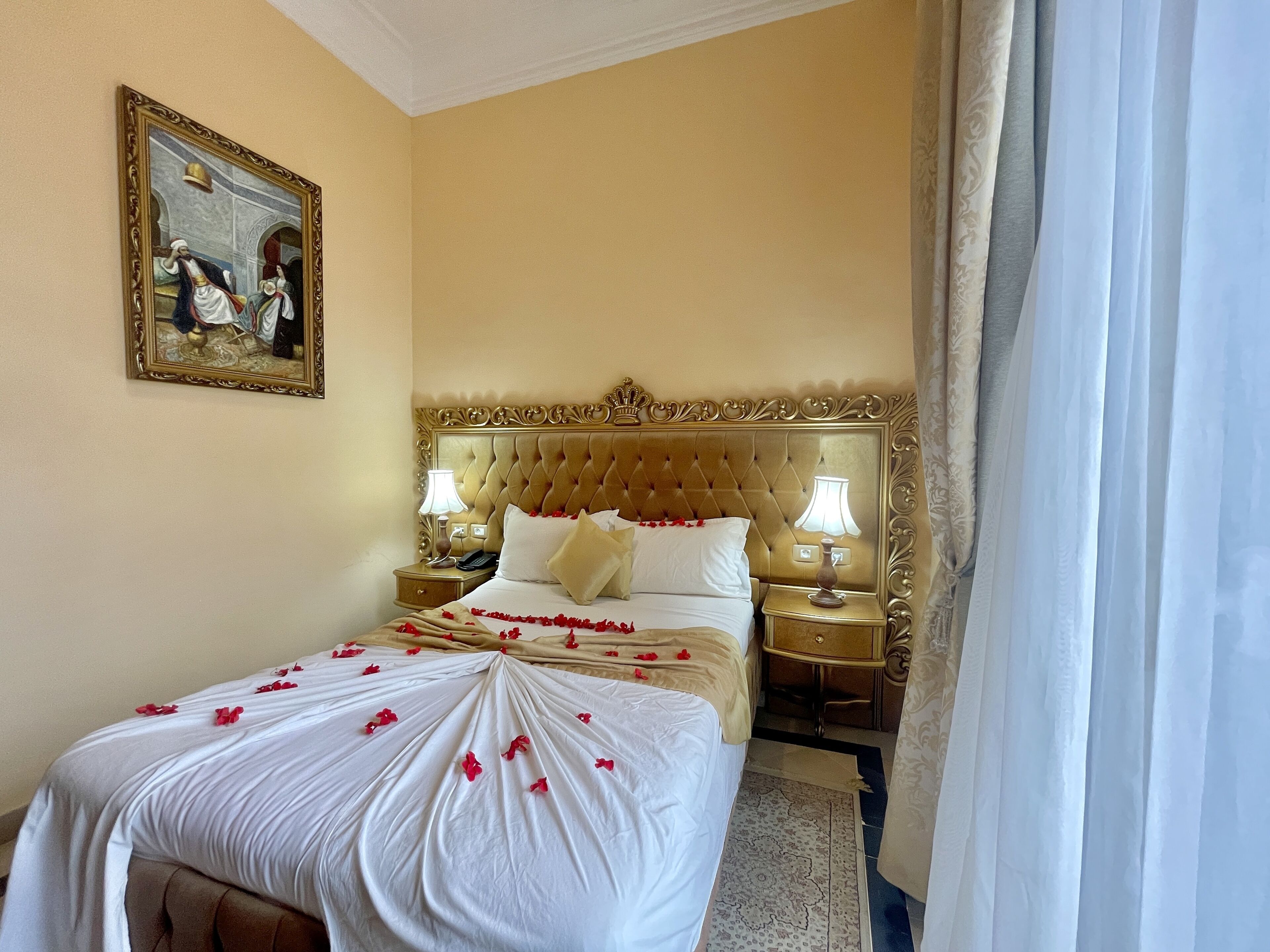 Quarto de hotel com uma cama grande, duas mesas de cabeceira, um espelho com moldura dourada e um quadro na parede.