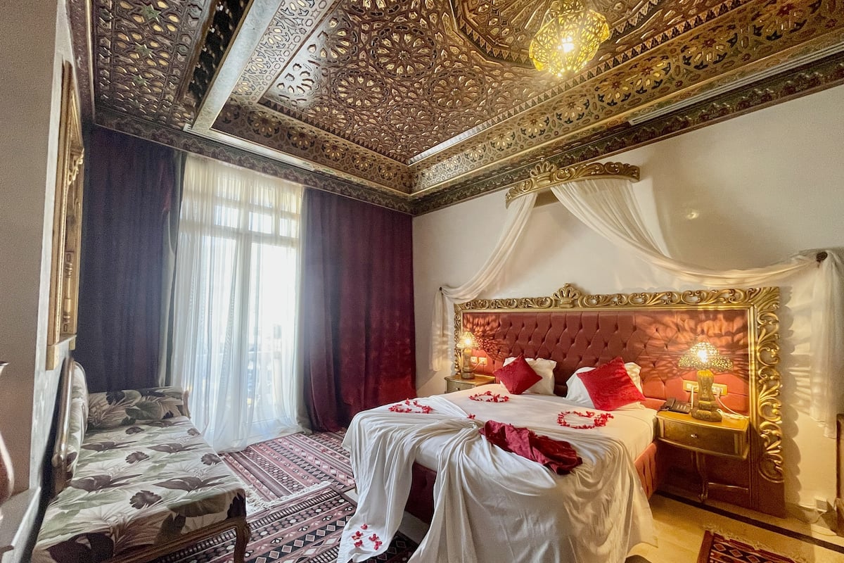 A luxurious bedroom with an ornate ceiling, a large bed with red pillows, and a chandelier.