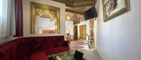 A room with a red sofa, a laptop on a glass table, ornate ceiling, and a painting on the wall.