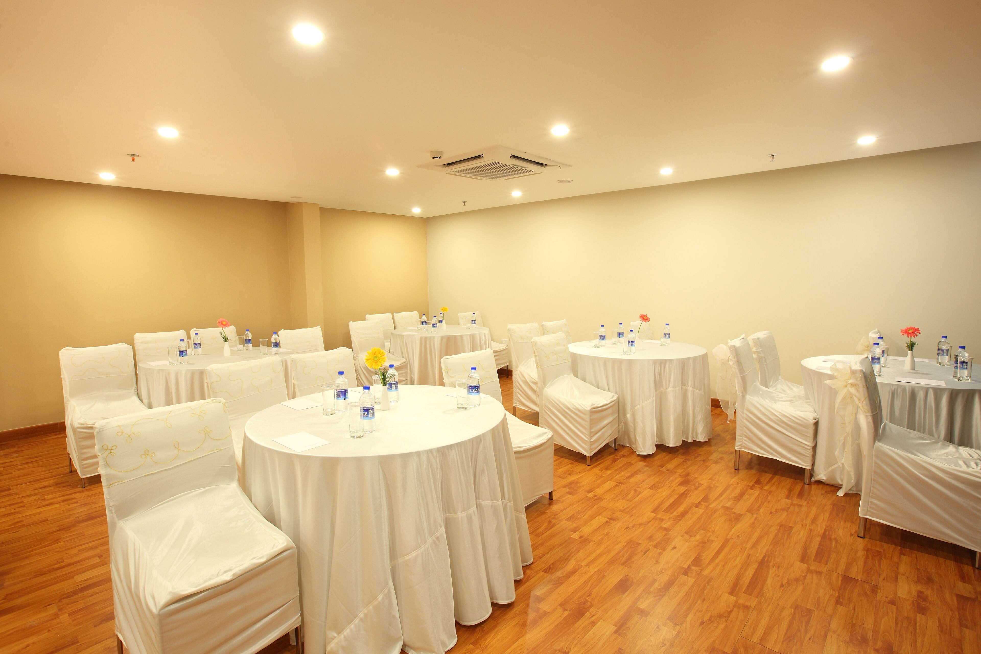 Un salón de conferencias con mesas redondas y sillas blancas, equipado con botellas de agua y pequeños arreglos florales.