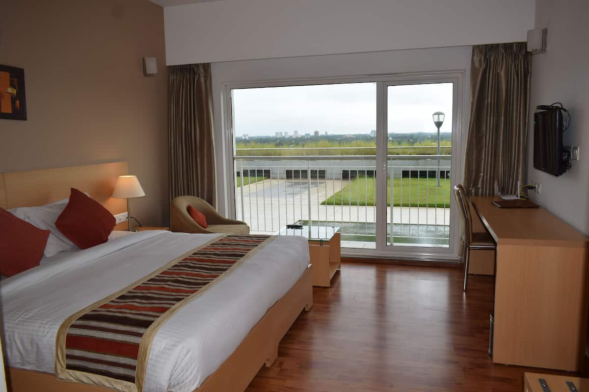 A hotel room with a large bed, a desk, a chair, and a view of a grassy area through a sliding door.