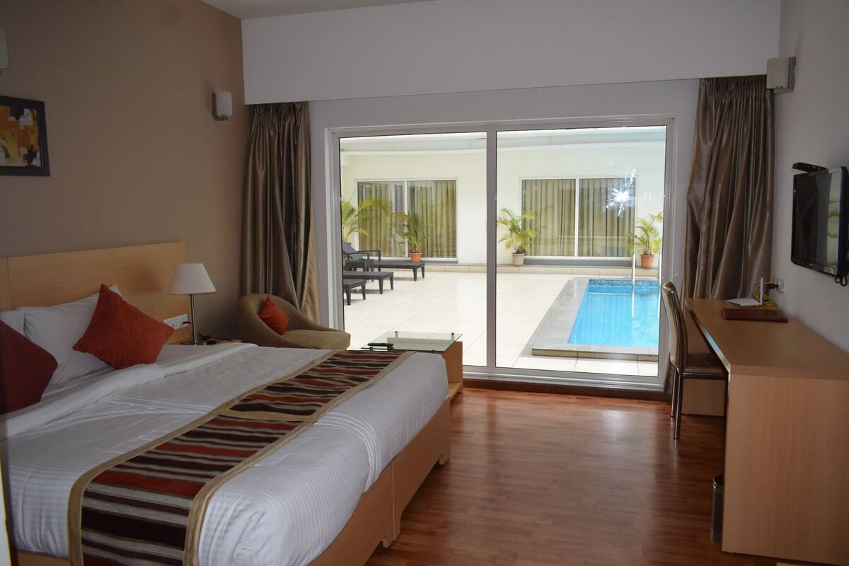 A hotel room with a large bed, a desk, a chair, a TV, and a view of a pool and outdoor seating area.