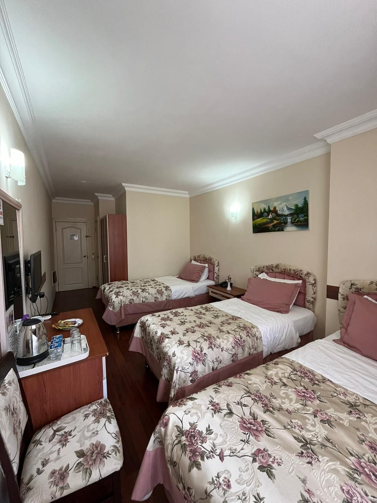 A hotel room with three beds, a desk with a kettle and glasses, and a painting on the wall.