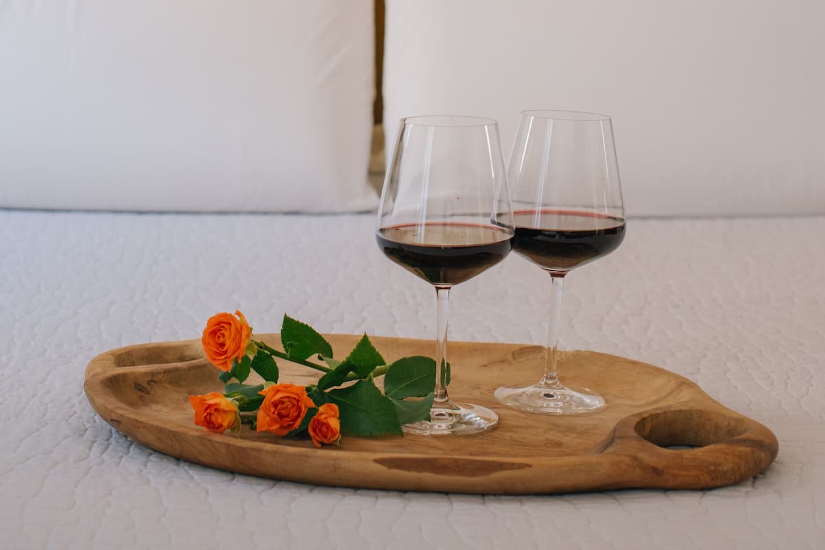 A bed with white and yellow bedding, two wine glasses on a wooden tray with roses, and a framed artwork on the wall.