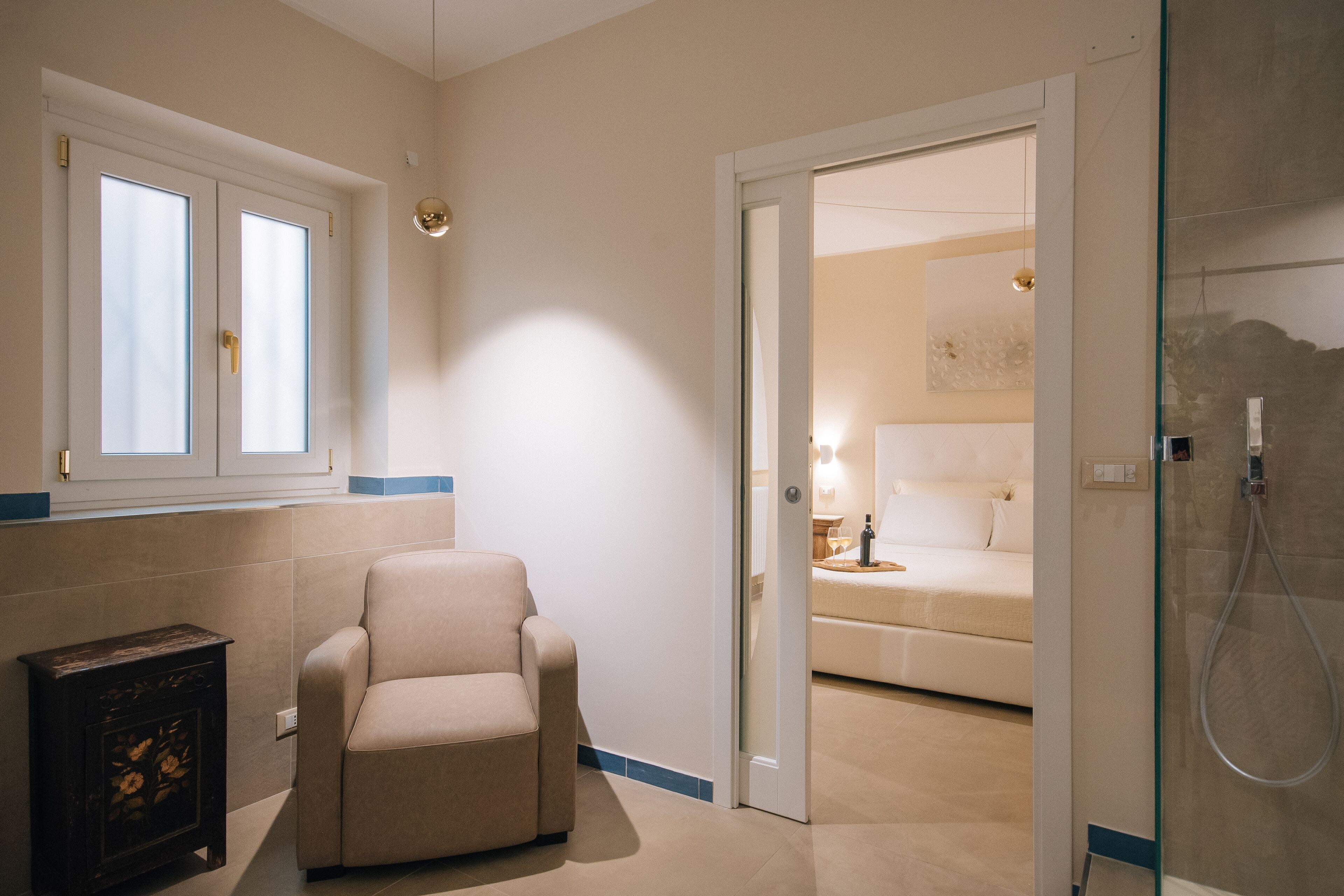 Une chambre d’hôtel avec un fauteuil beige, un lit avec une literie blanche, une douche en verre et une petite armoire ornée de motifs floraux.