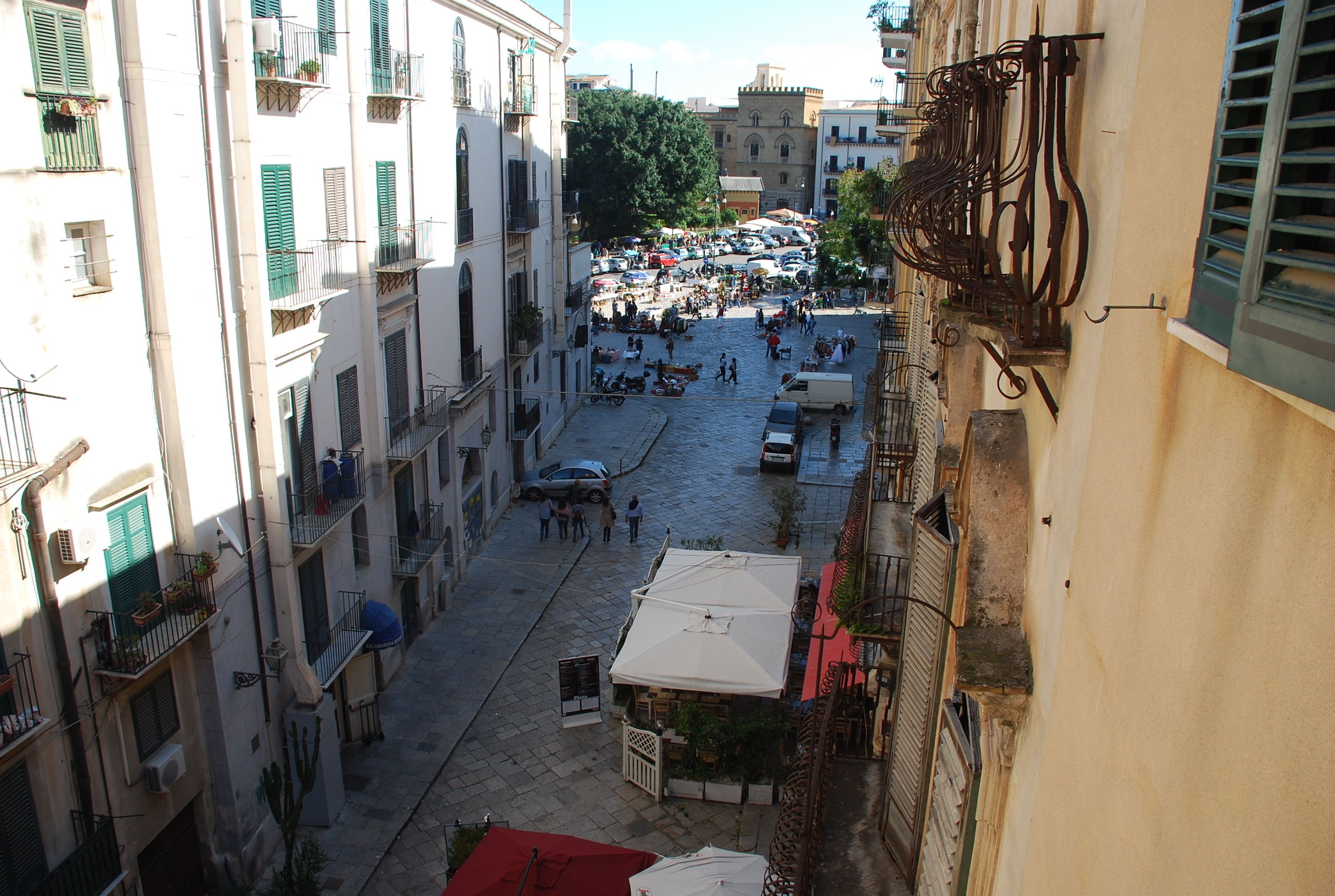 A narrow street with buildings on both sides, outdoor seating areas, and a few parked cars.