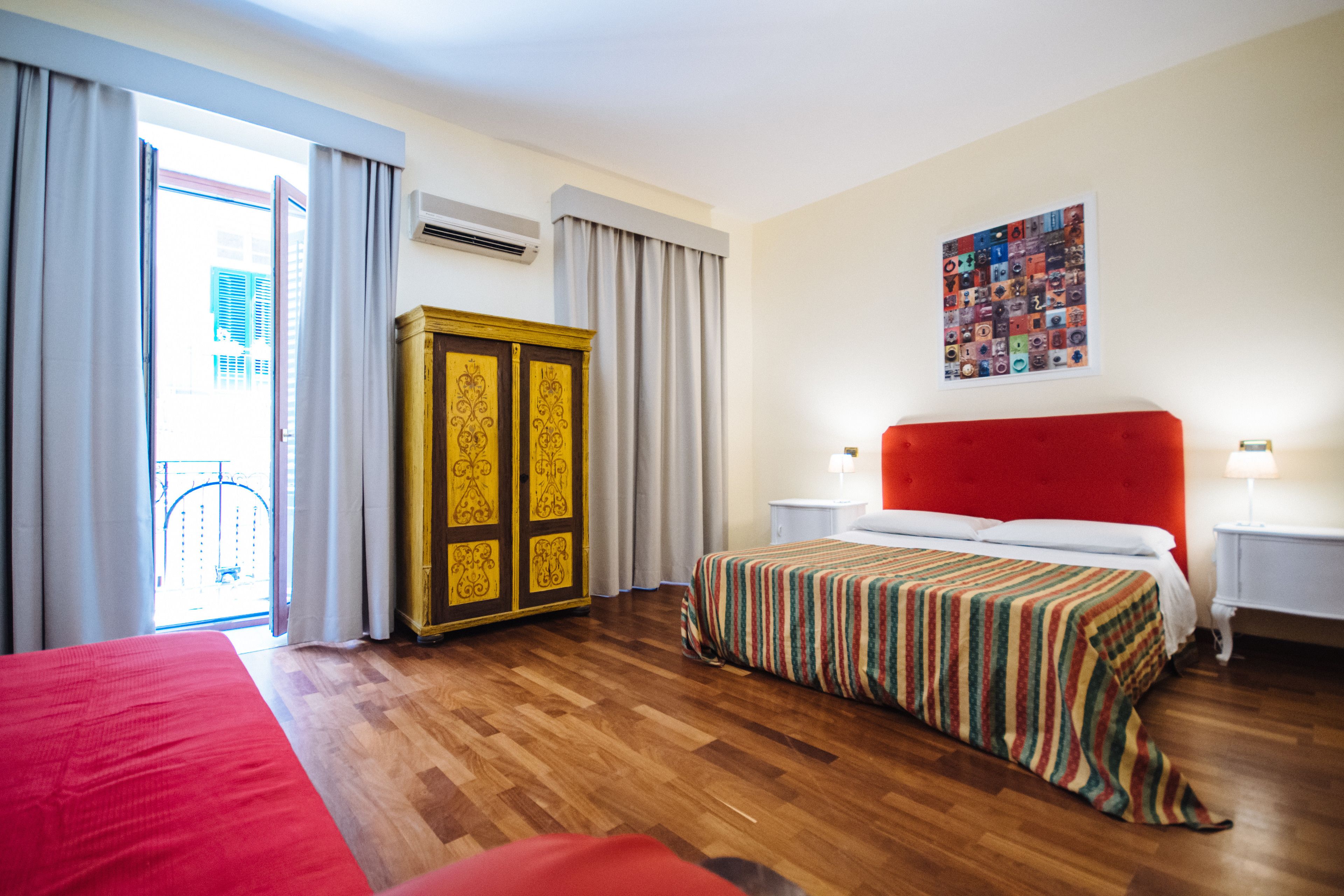 A hotel room with a wooden floor, a red bed, a large wardrobe, a red headboard, a nightstand with a lamp, and a window with curtains.