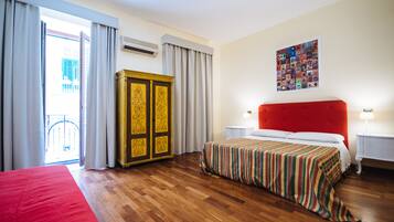 Une chambre d’hôtel avec un plancher en bois, un lit rouge, une grande armoire, une tête de lit rouge, une table de chevet avec une lampe et une fenêtre avec des rideaux.