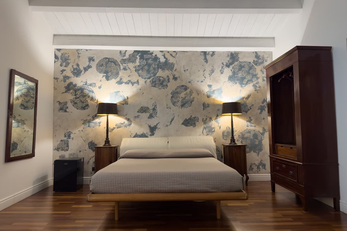 A bedroom with a large bed, two bedside tables with lamps, a wardrobe, and a patterned wallpaper.